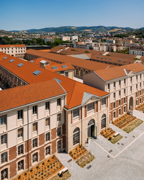 Aerial view of terracotta-roofed buildings and courtyard at Cité du Design in Saint-Étienne with city and mountains in background.