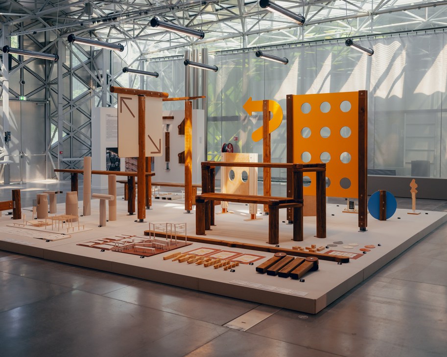 Exhibition display at La Platine featuring wooden furniture structures, orange geometric panels, and architectural models in a modern gallery space.