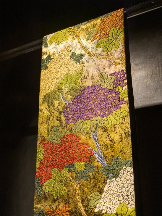Illuminated textile panel featuring embroidered red, purple, and white floral clusters on a distressed gold background by Genbei Yamaguchi.