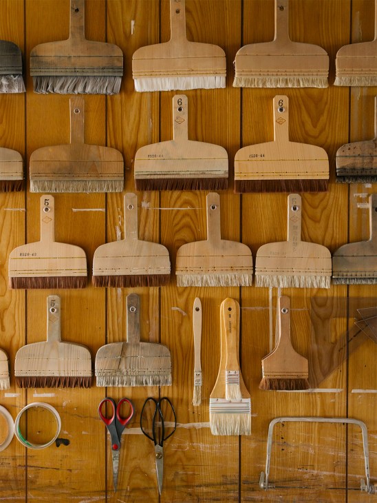 Collection of wooden-handled paintbrushes with natural bristles arranged on a wooden pegboard wall.