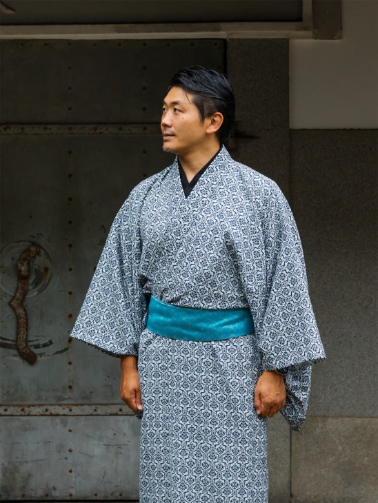 Man wearing blue patterned kimono with turquoise obi standing against textured wall.