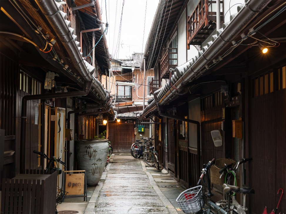 Traditional wooden buildings line a narrow Kyoto street with bicycles parked along the alley.
