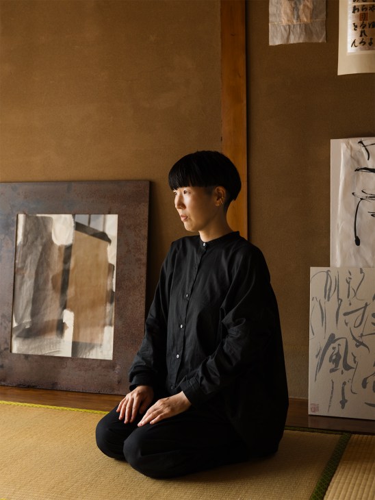 Calligrapher Mikiko Kayama sitting in seiza position in her studio surrounded by artworks on tan walls.