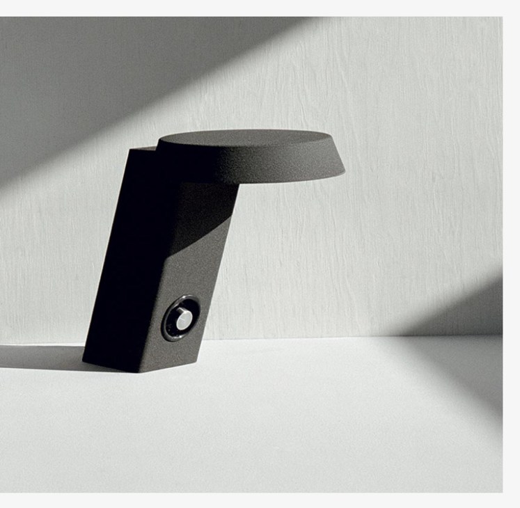 Modern minimalist black and gray sculptural object on white surface with diagonal shadow.