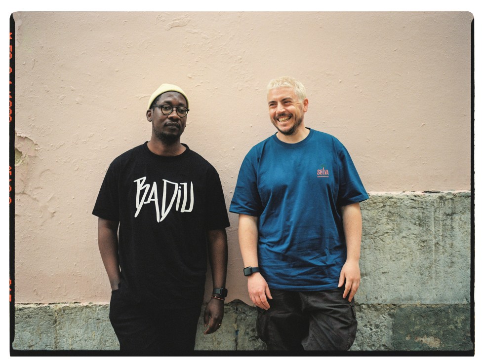Two men stand smiling against a weathered beige wall. The man on the left wears glasses and a black t-shirt, while the man on the right wears a blue t-shirt.