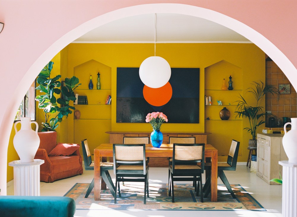 Dining room with yellow walls, wooden table, wicker chairs, pendant light, and colorful artwork