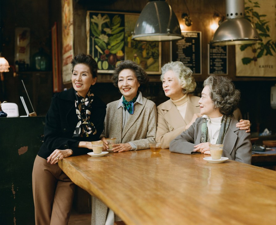Four elderly women sitting together at a wooden table in a cafe, smiling and drinking coffee