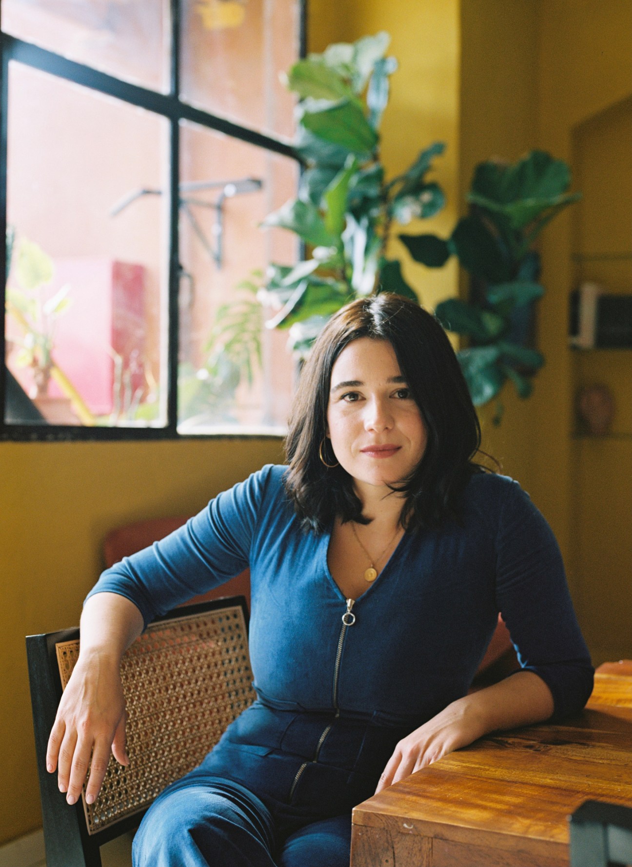 Sara Andújar sits at a wooden table in a colorful interior space with plants and windows
