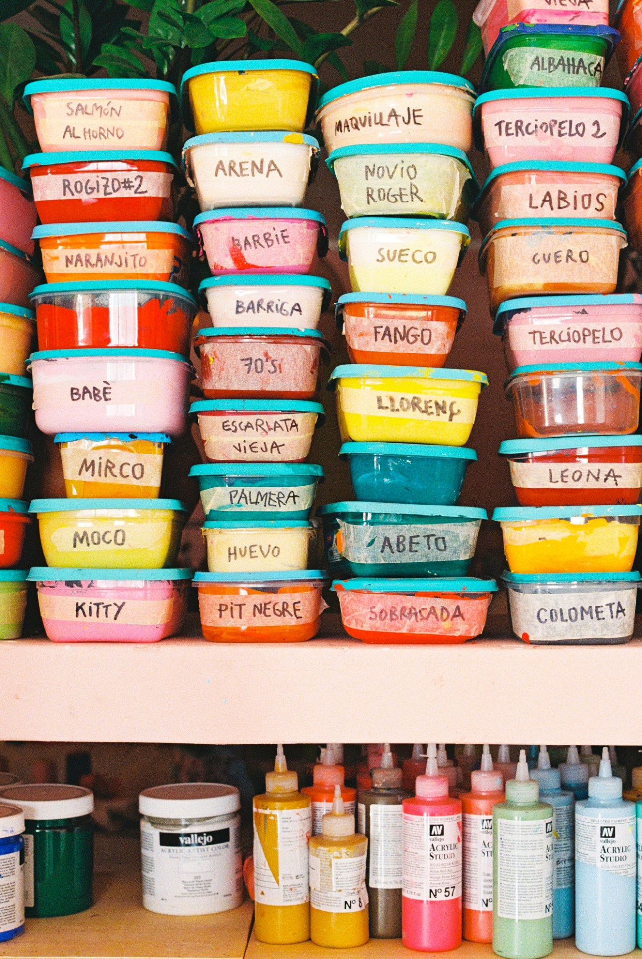 Stacked containers of colorful acrylic paint with handwritten Spanish labels in an artist's studio