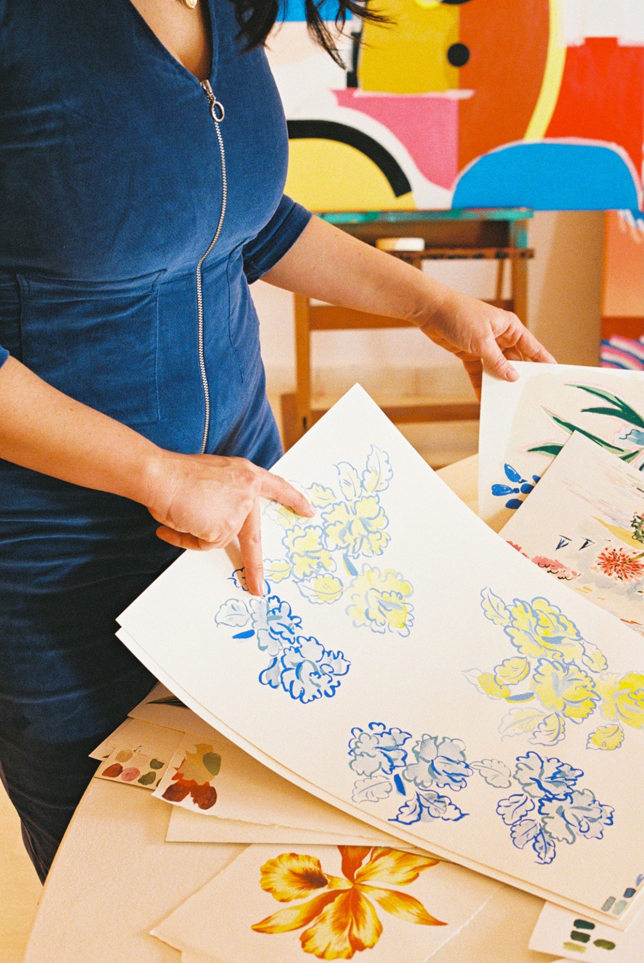 Woman in blue top pointing at floral sketches with blue and yellow watercolor paintings spread on wooden table