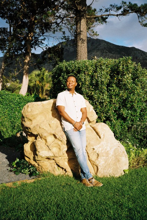 Beks Ndlovu, CEO of African Bush Camps, stands in front of a large rock formation in a lush garden setting with mountains in the background.