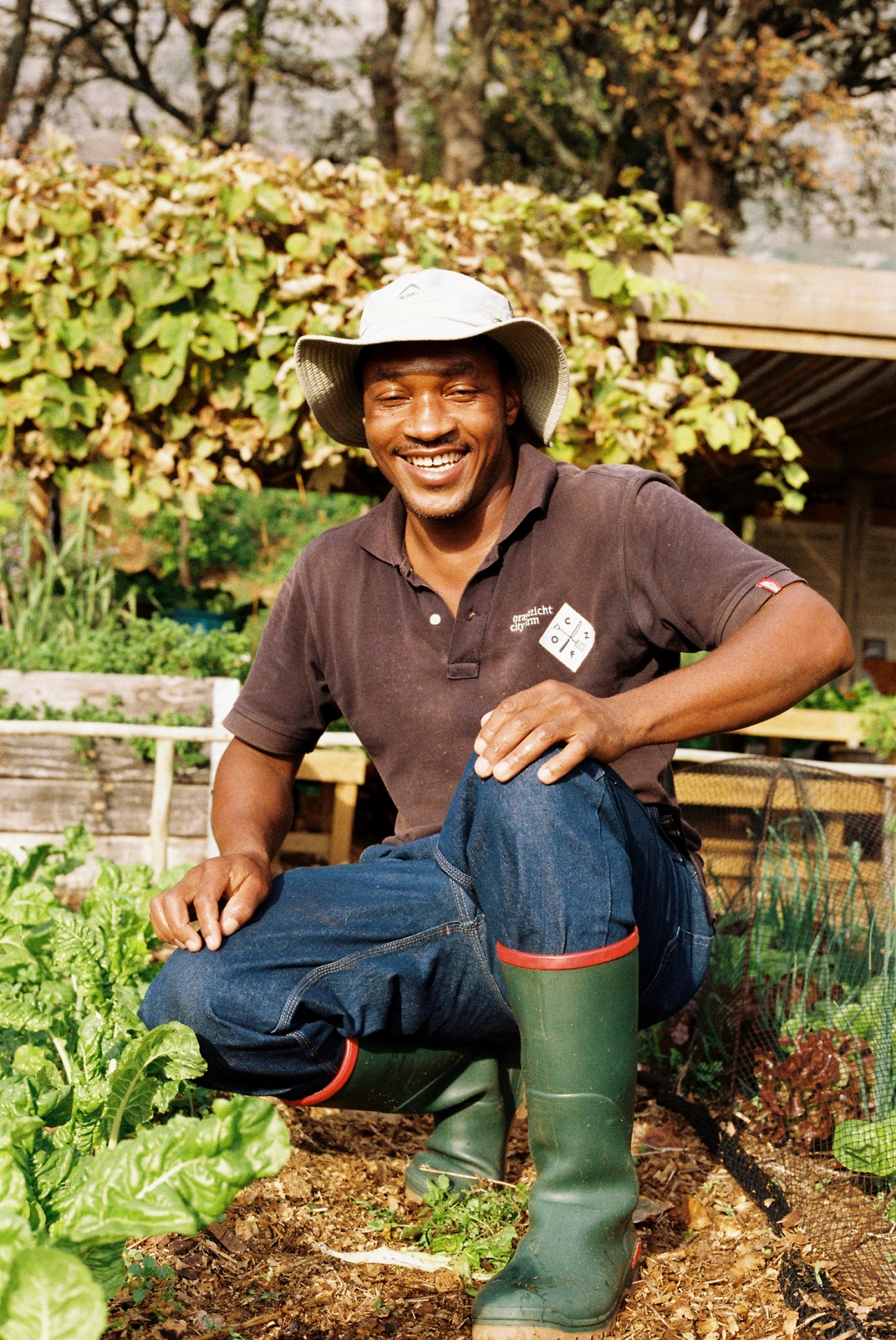 Kenneth Masinga smiling while kneeling in a garden wearing a hat, brown polo shirt, jeans and green wellington boots