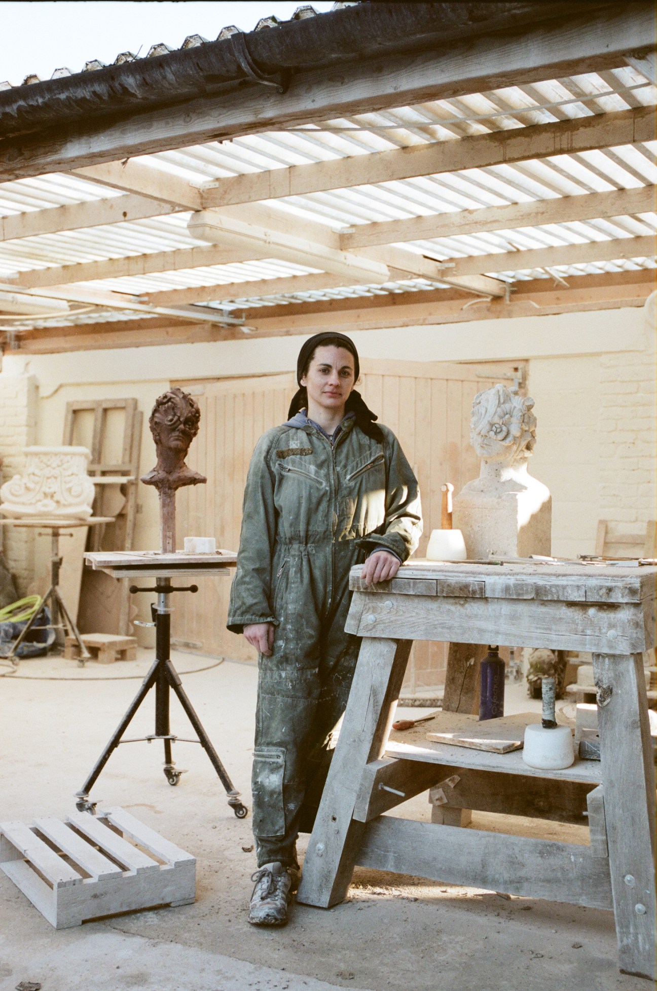 Anna Rubincam stands in her stone-carving workshop wearing work clothes beside carved sculptures and tools