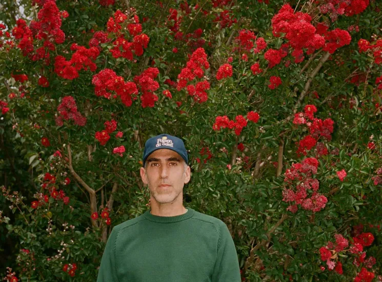Brendon Babenzien wearing a green sweater and baseball cap standing in front of red flowering trees