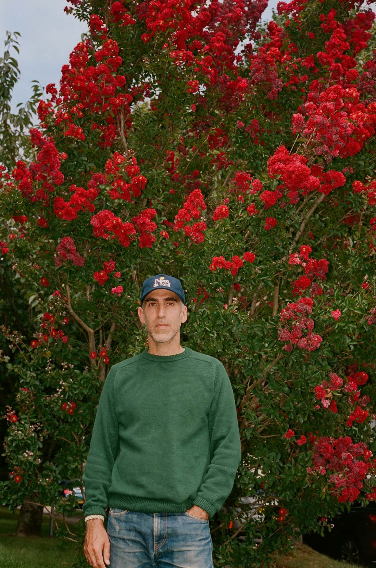 Brendon Babenzien wearing a green sweater and baseball cap standing in front of red flowering trees