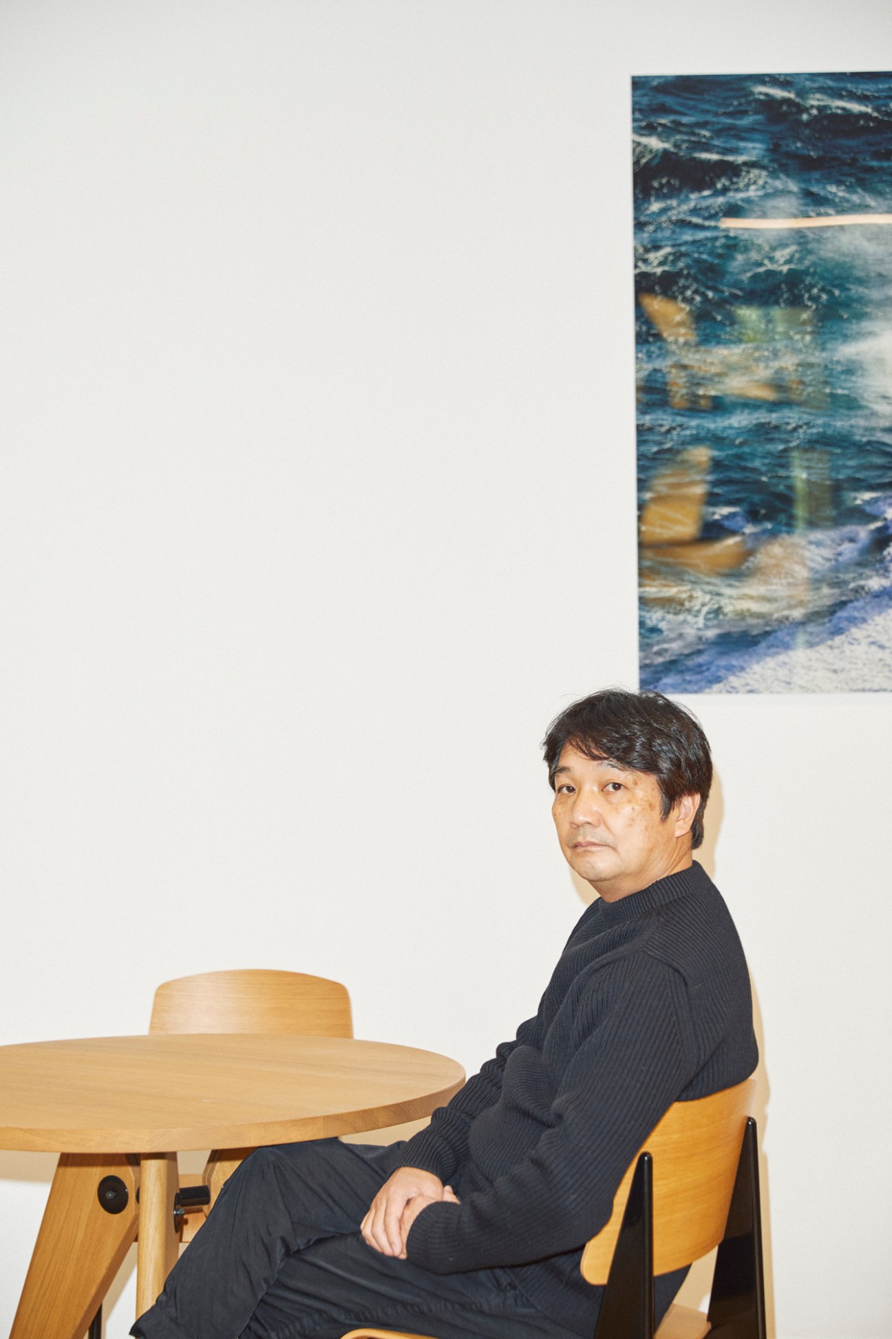 Takao Watanabe sitting at wooden table wearing dark sweater with ocean artwork on wall behind