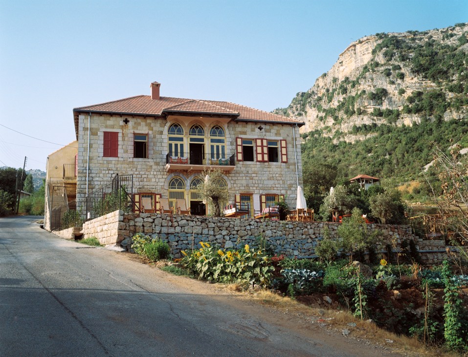 A traditional Lebanese stone house with yellow and red shutters nestled against dramatic mountain cliffs.