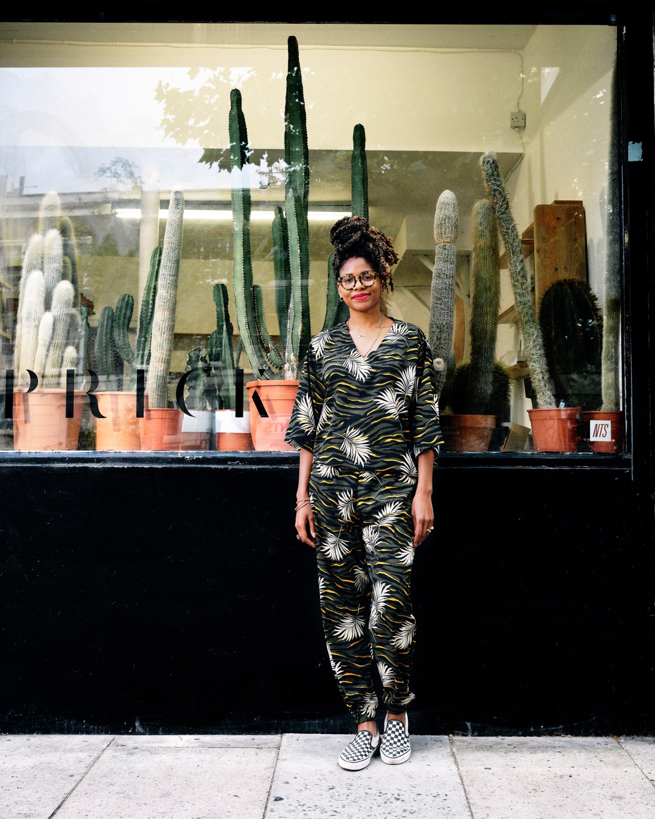 Gynelle Leon standing in front of her London plant shop Prick, wearing a palm print jumpsuit with cacti visible in the window