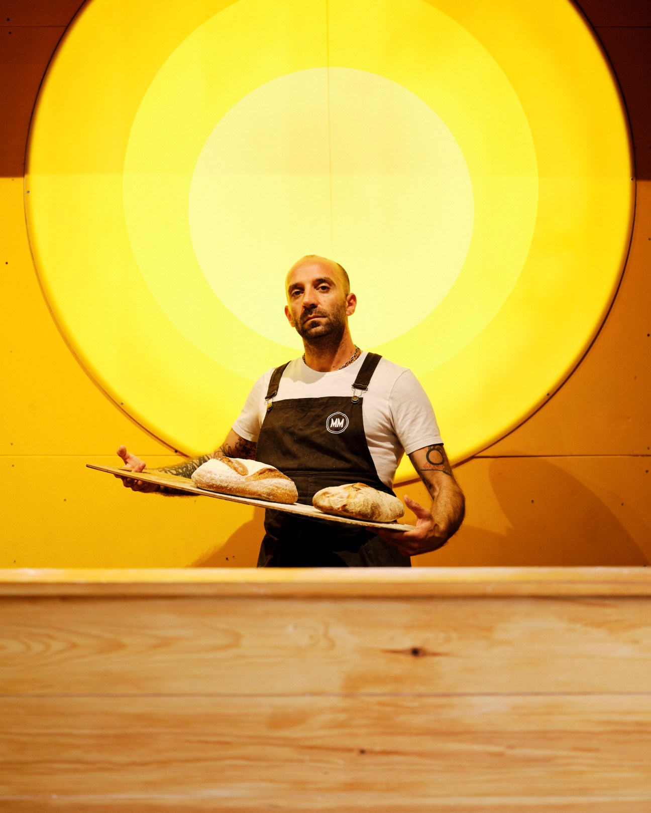 Baker with tattoos holding wooden board with fresh bread loaves in front of large circular yellow light