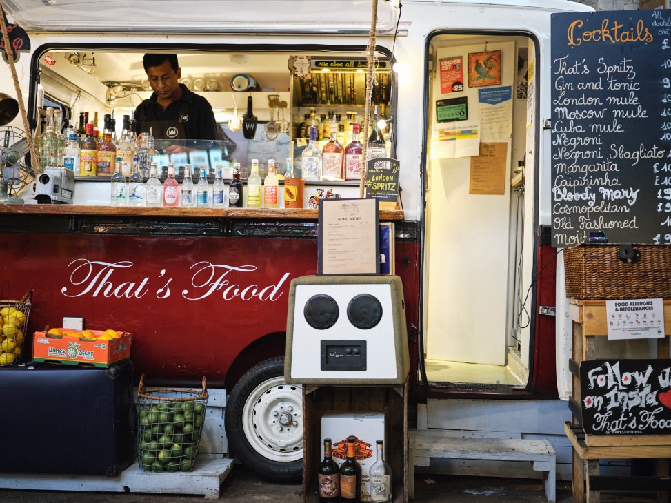 Mobile food truck with red and white exterior displaying 'That's Food' branding and cocktail menu at market