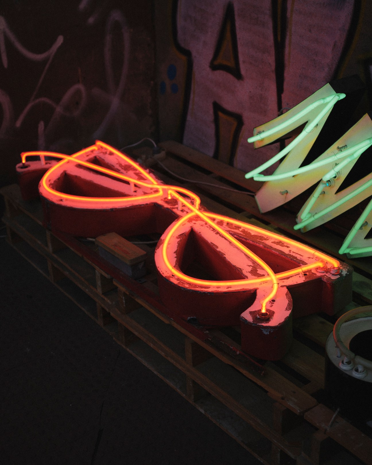 Red and orange neon sunglasses sign with green neon letters on wooden workbench in workshop