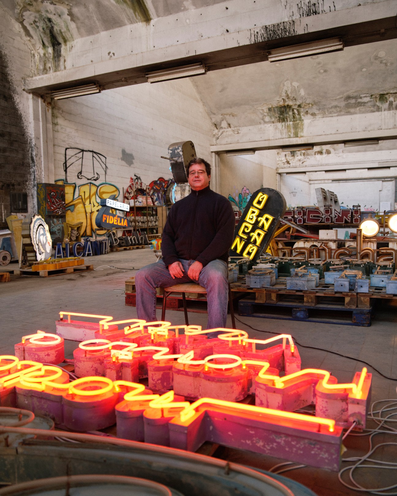 Paulo Barata sits in a warehouse surrounded by colorful vintage neon signs and letters