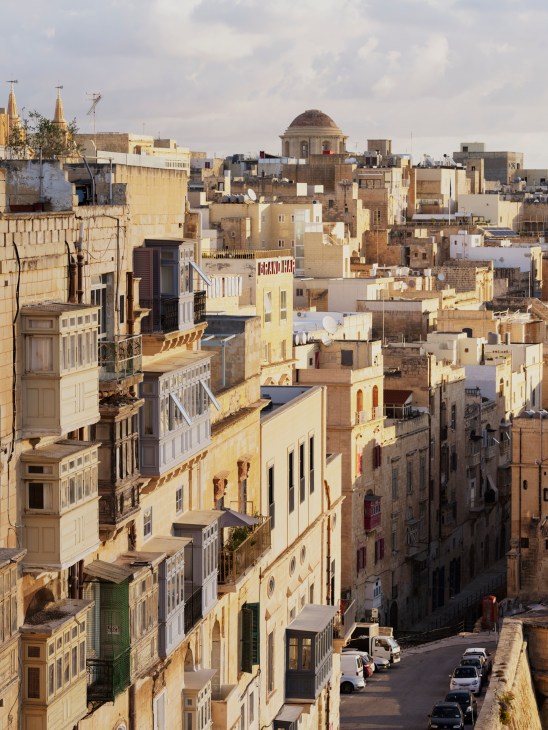 Aerial view of Valletta's densely packed golden limestone buildings with domed structures and seafront in background.