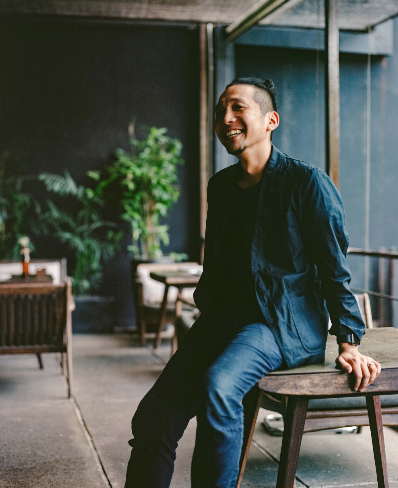 Man in denim shirt sitting on wooden chair in modern office with plants and windows.