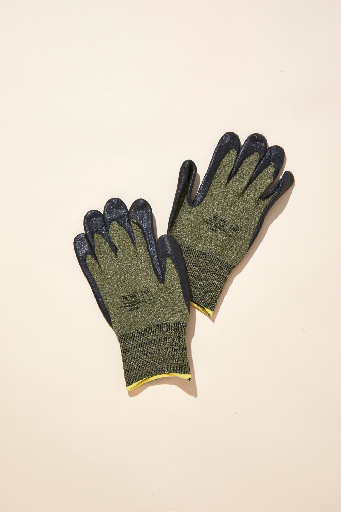 A pair of olive green work gloves with dark fingertips and yellow cuff trim laid on a beige surface