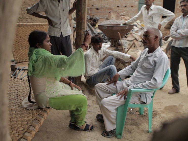 A woman in green traditional dress sits facing an elderly man in white clothing in a rural courtyard setting