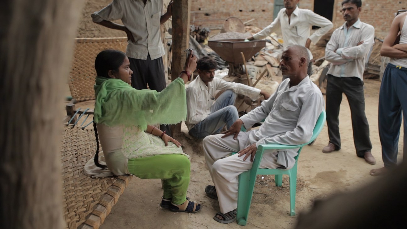 A woman in green traditional dress sits facing an elderly man in white clothing in a rural courtyard setting