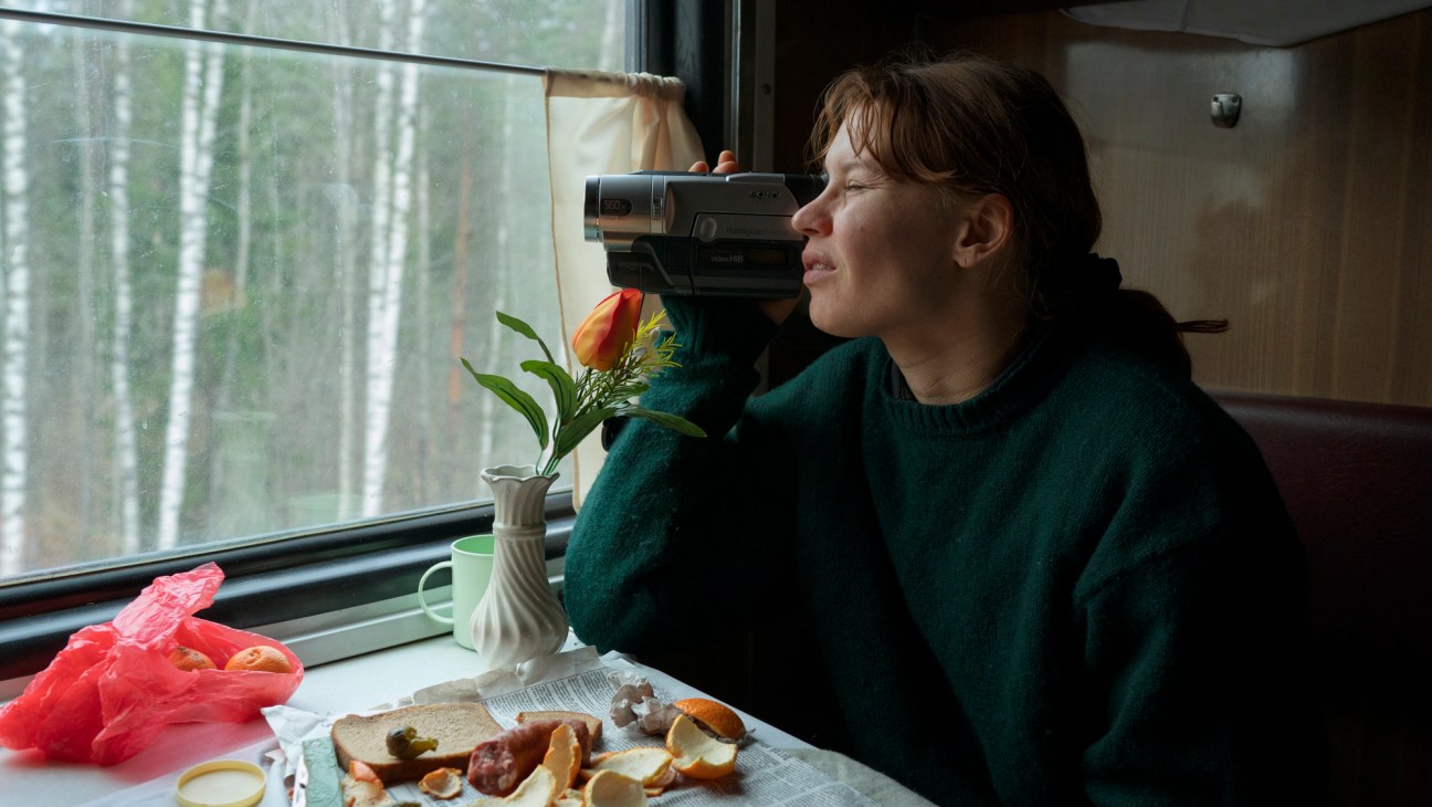 A woman in a green sweater looks through a video camera while sitting at a train table with food and flowers