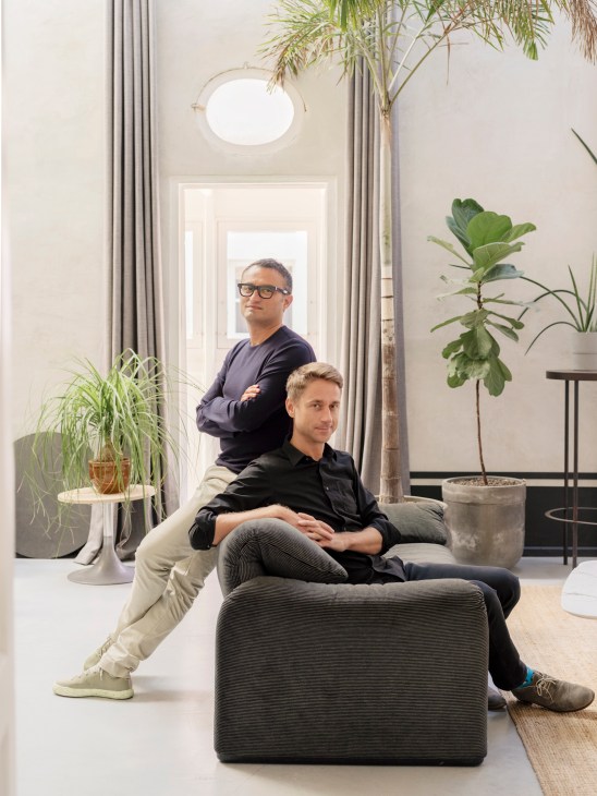 Architects Chris Briffa and Leroy Merks pose in a bright, minimalist interior space with plants and natural light.