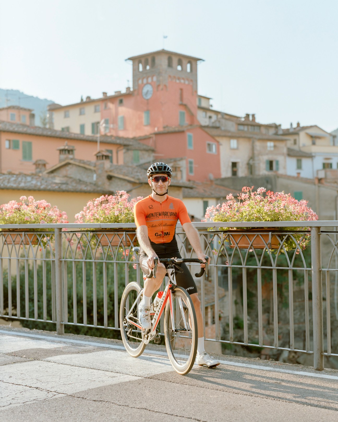 Cyclist in orange jersey with helmet standing with bike on bridge overlooking historic Italian hillside town