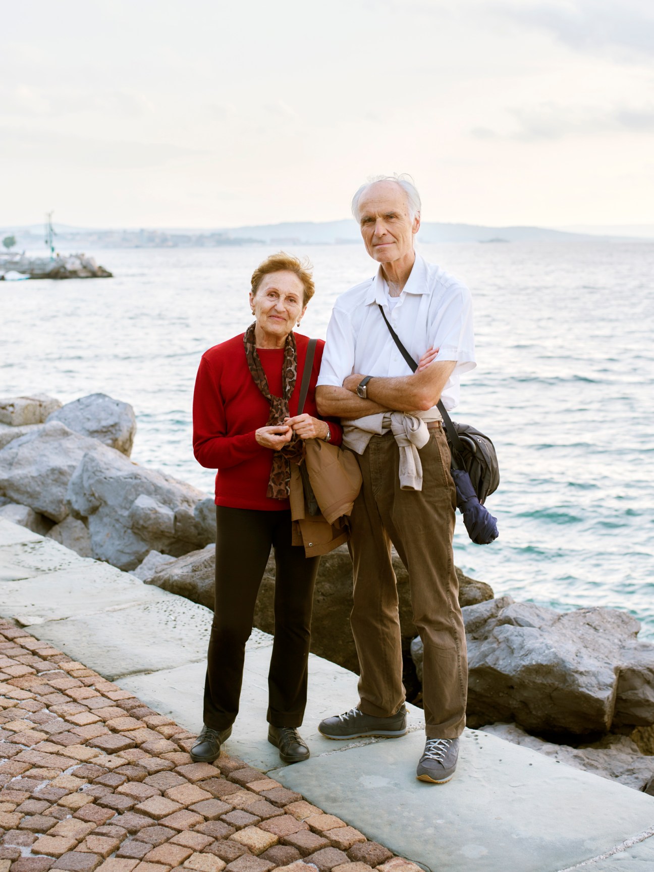 Two people standing together on a waterfront promenade with calm sea and distant hills in background.