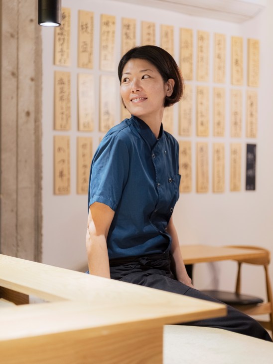 Chef Miyuki Vinet stands in her restaurant with calligraphy artwork displayed on the wall behind her.