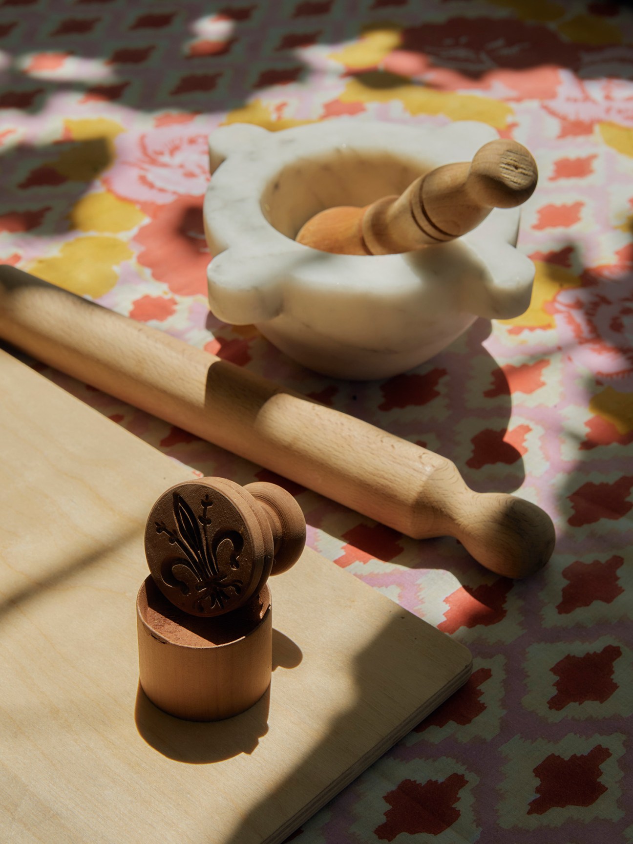 Wooden kitchen tools including a rolling pin, decorative wooden stamp with leaf pattern, and white marble mortar and pestle on a floral patterned tablecloth