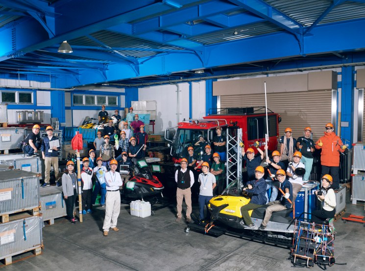 A large group of workers in hard hats pose together inside an industrial warehouse with vehicles and equipment around them.