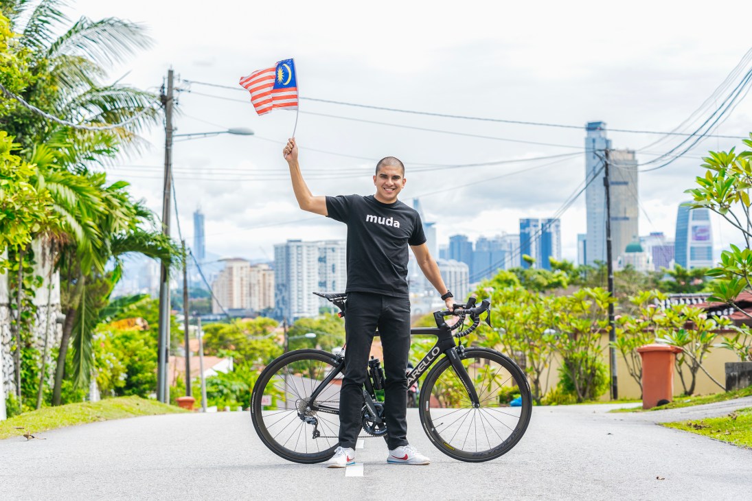 Syed Saddiq stands with his bicycle holding a Malaysian flag, wearing a black Muda t-shirt with city skyline behind