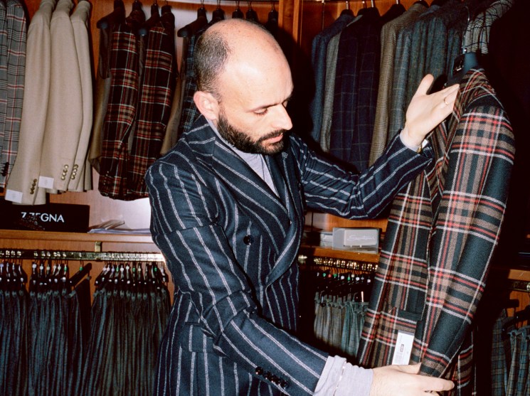A bearded man in a pinstripe suit browses tartan fabrics in a high-end menswear shop.