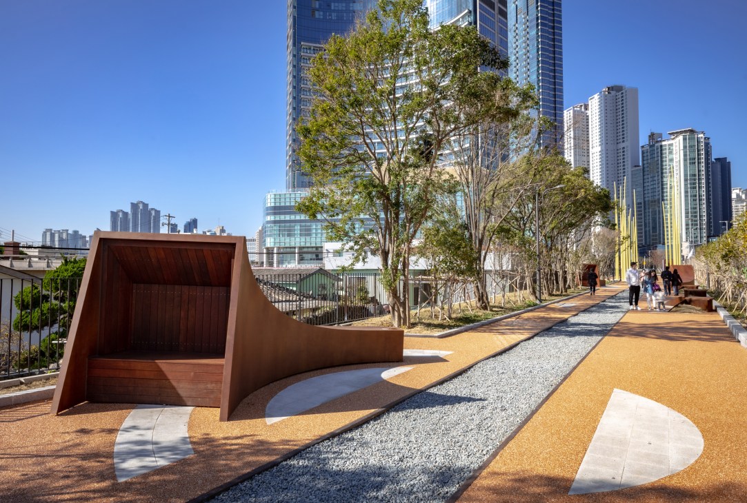 Modern sculptural wooden seating structure on colorful paved walkway in Haeundae Blueline park with city skyline