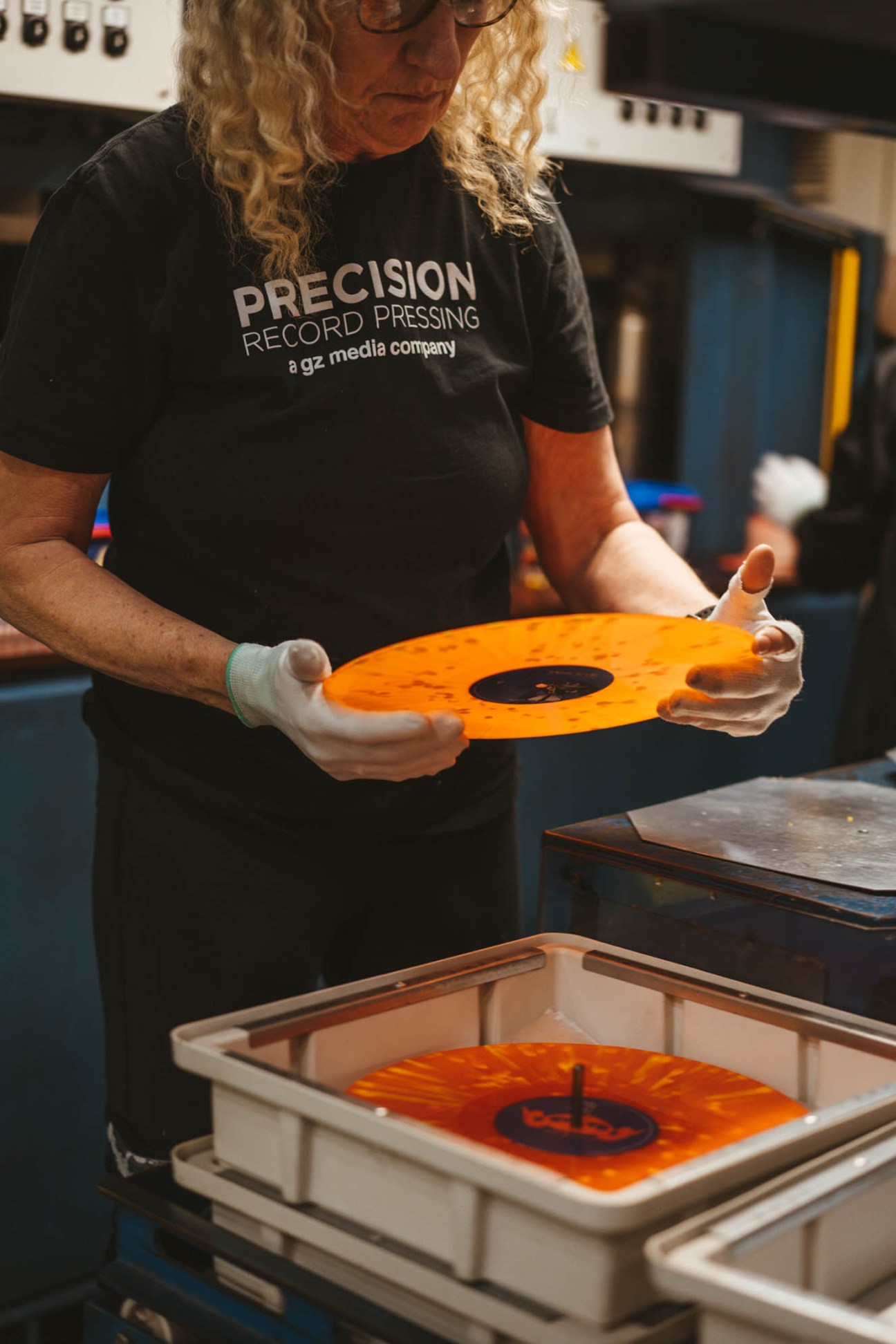 Person in Precision Record Pressing t-shirt holding bright orange vinyl record above cooling tray in production facility