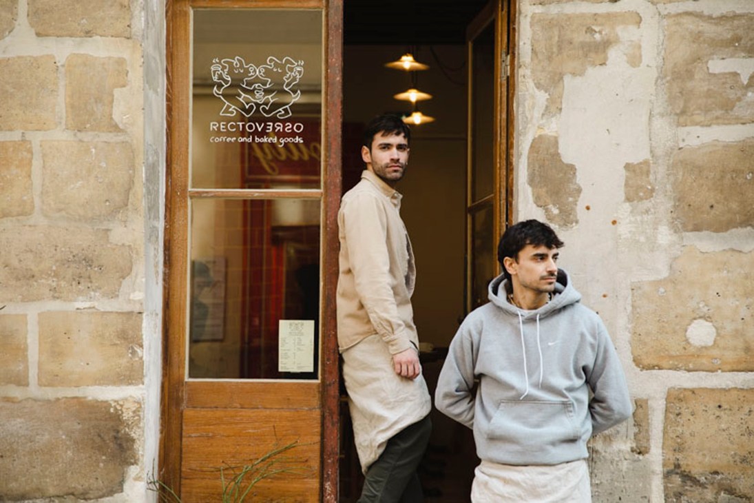 Two men standing outside Recto Verso cafe storefront with stone facade and glass door