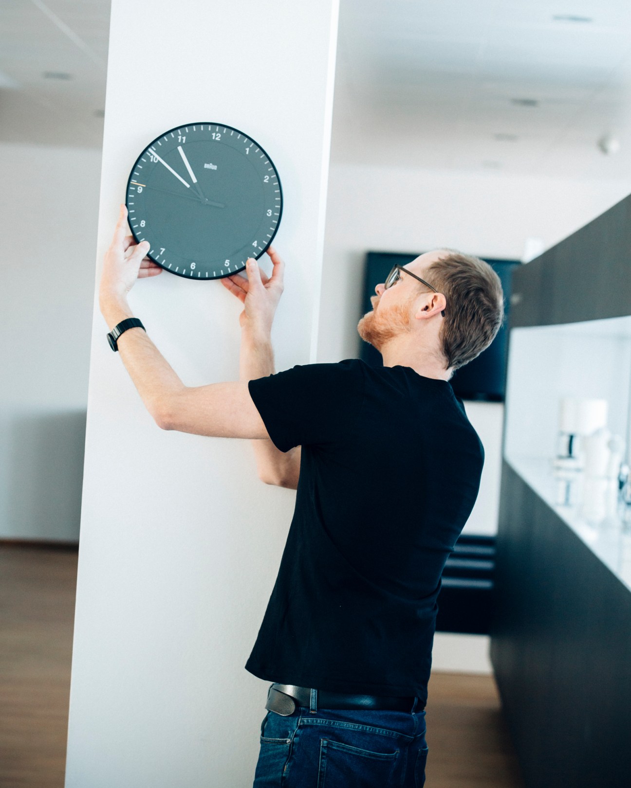 Man in black shirt mounting a dark wall clock on white pillar in modern interior