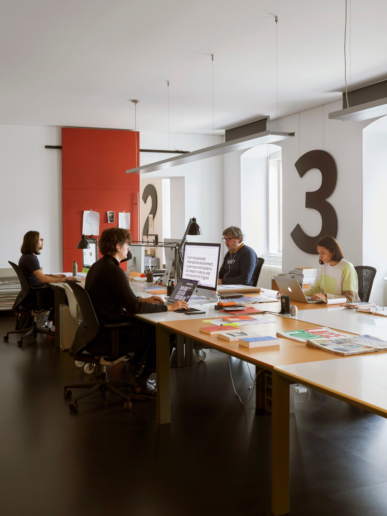 Interior of a modern office workspace with four people working at a wooden table with laptops and design materials.