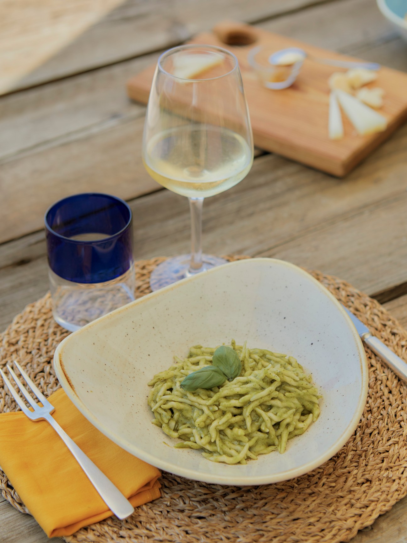 Bowl of green pesto pasta with basil garnish served with white wine and cheese board on woven placemat