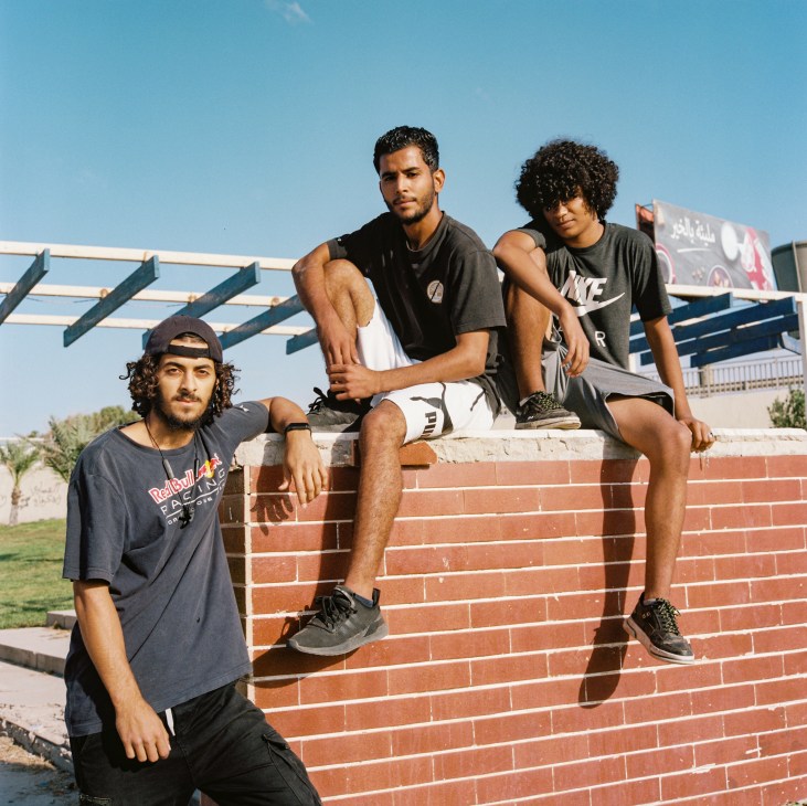 Three young men practicing parkour outdoors, one standing by a brick wall, two sitting on top