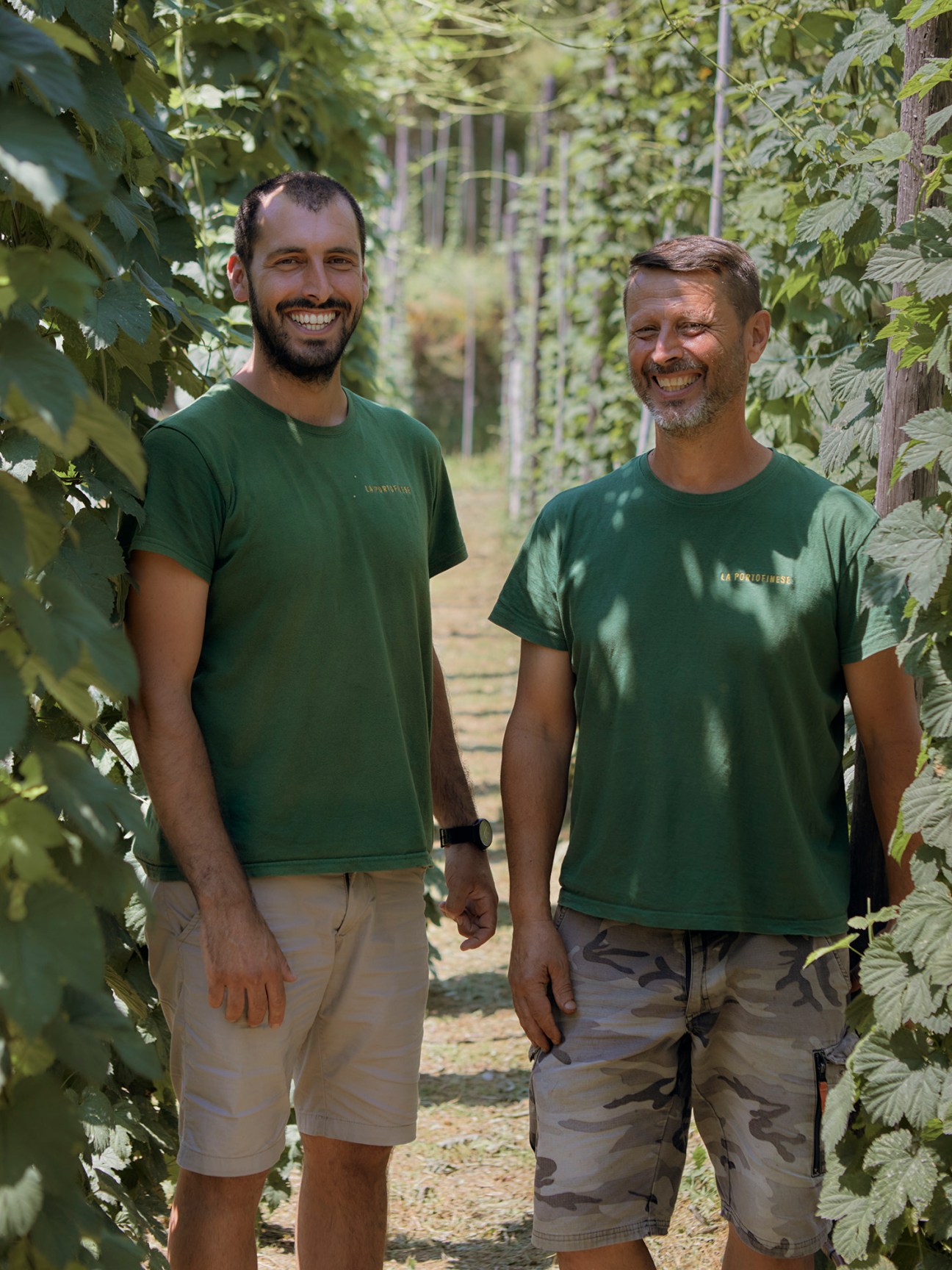 Two men in green t-shirts standing among leafy vine plants in a rural Italian agricultural setting