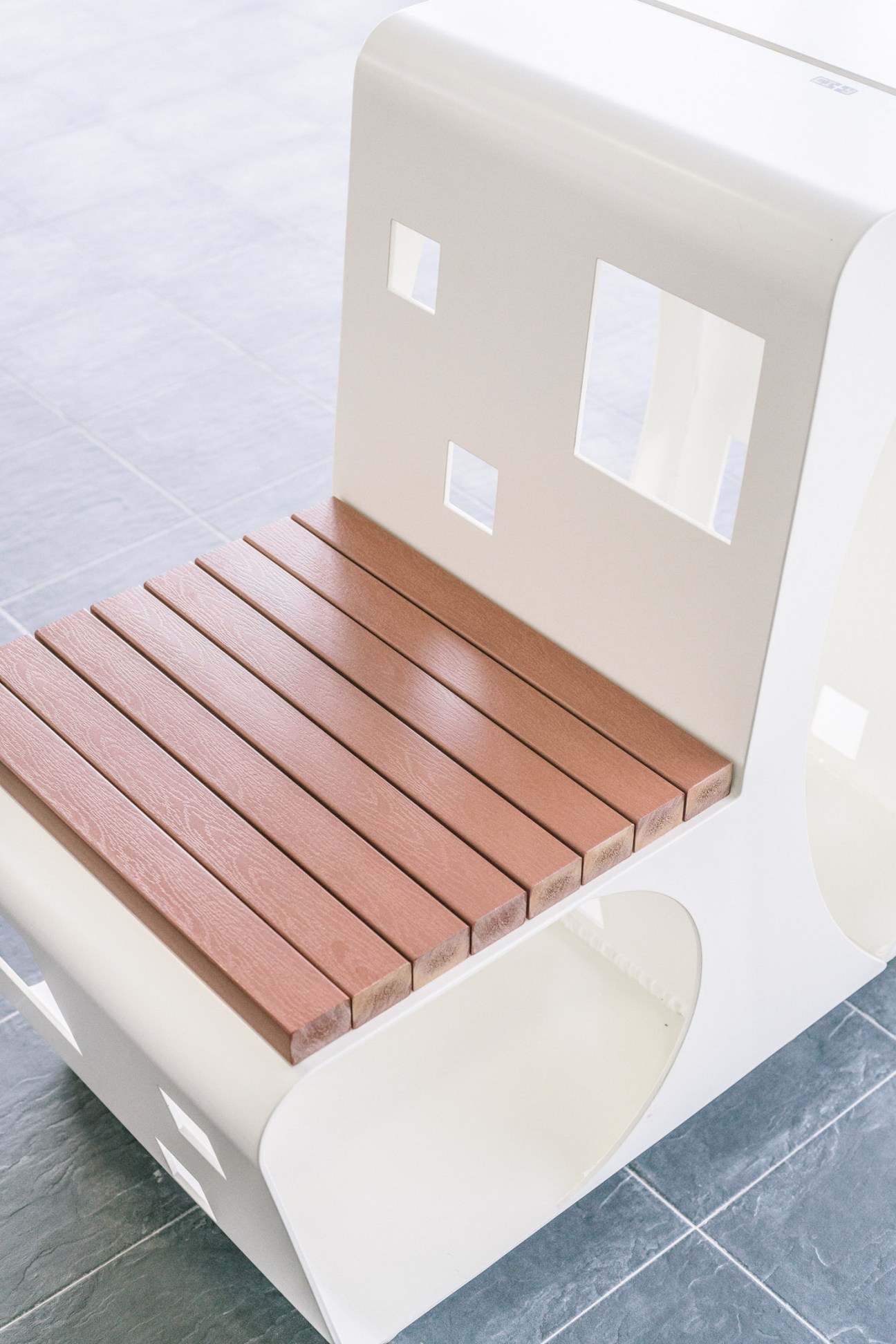 Program public bench with white top and wooden slats, featuring small window openings in the backrest.