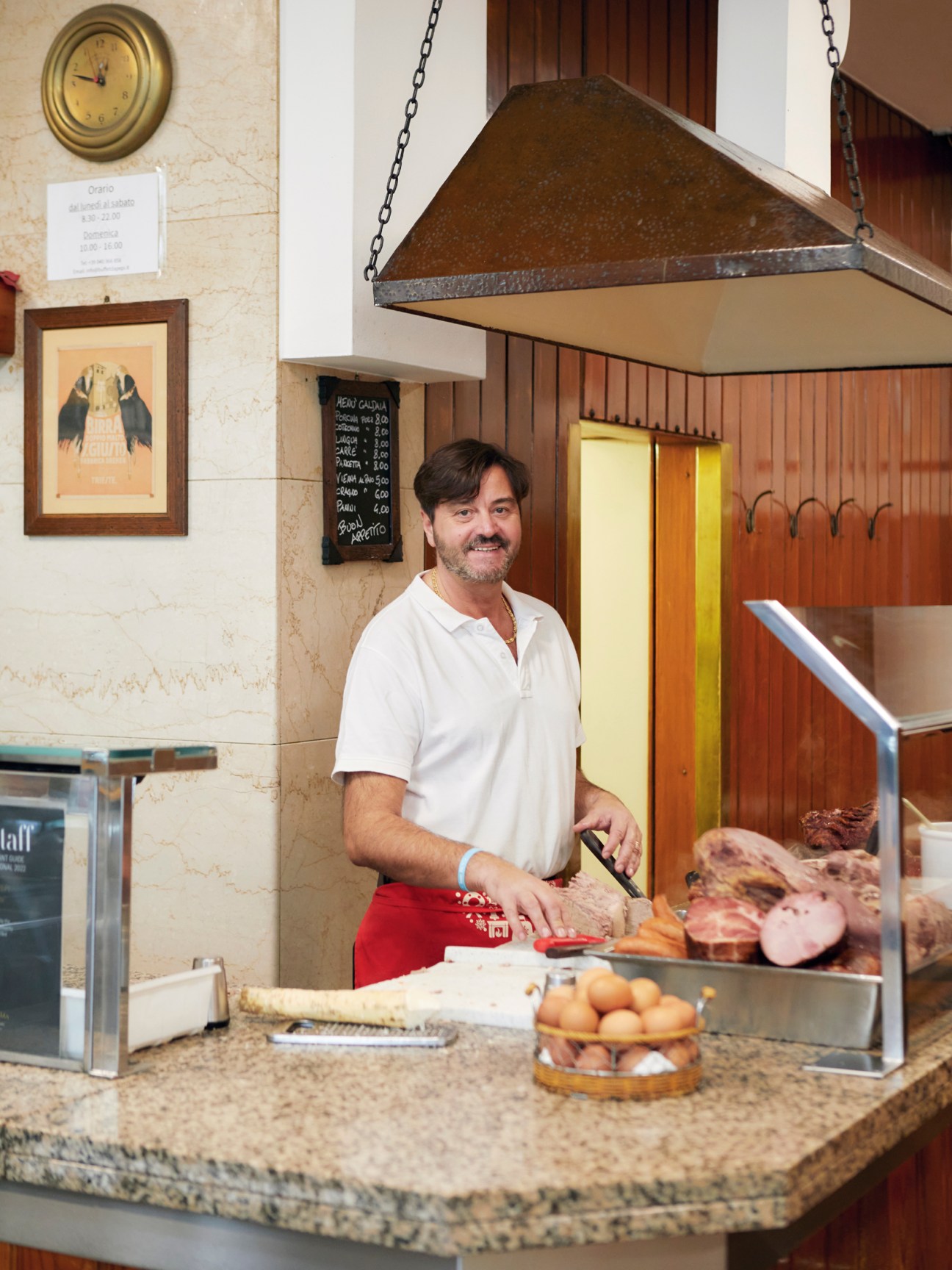Man in white shirt and red apron at a delicatessen counter with cured meats and products on display.